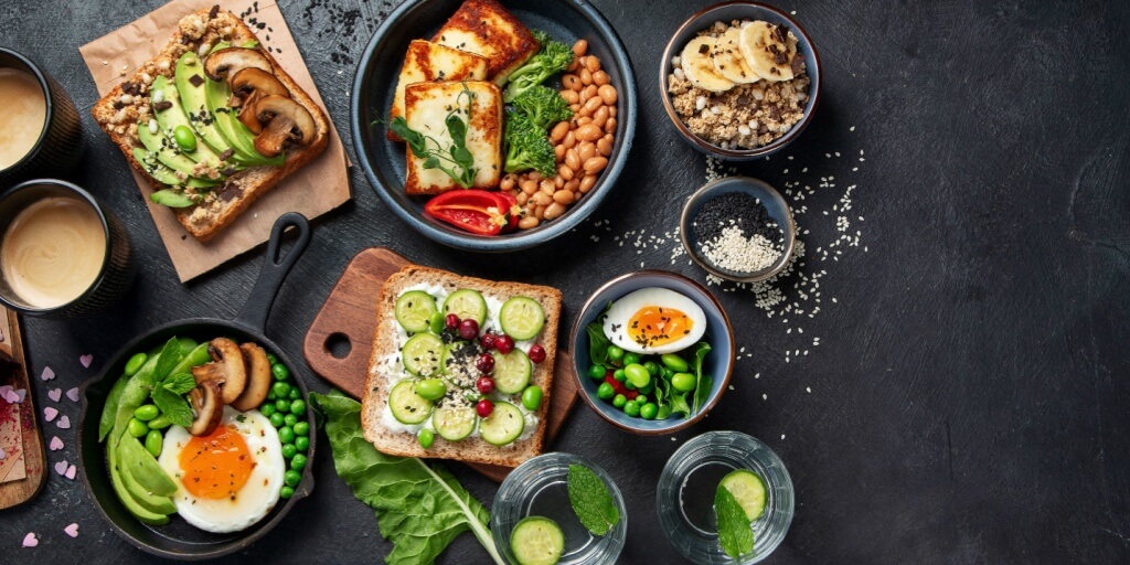 Ett färgglatt och nyttigt frukostbord med varierade rätter som avokado- och svamptoast, stekt ägg, sallad med gröna ärtor, och skivor av halloumi. I bakgrunden finns även en skål med gröt toppad med banan och en kopp kaffe. Allt serveras på en rustik mörk yta.